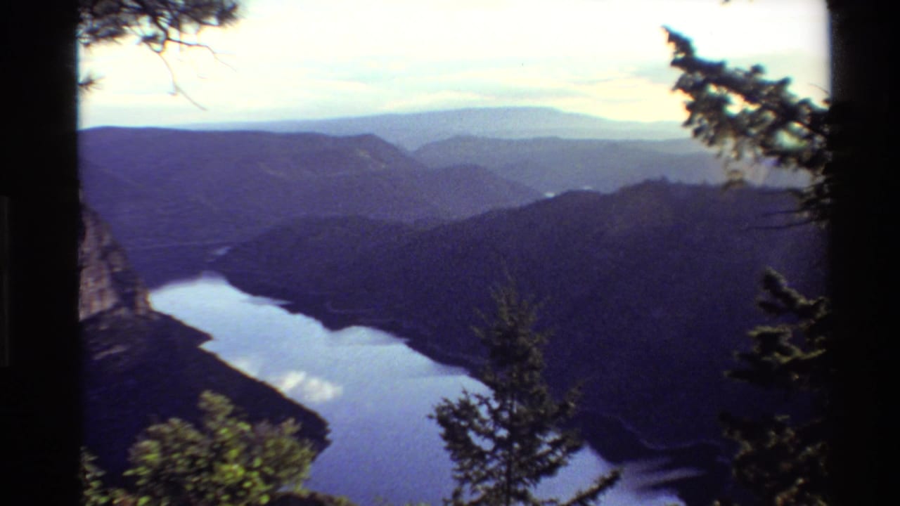 1980 Western USA 8mm footage frame with a lake curving through a forested valley, distant blue ridges, and trees along the overlook.