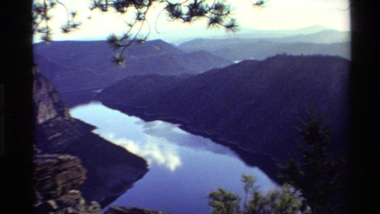 1980 Western USA 8mm frame showing a reflective river winding below mountains, with pine branches and rocky edges framing the view.