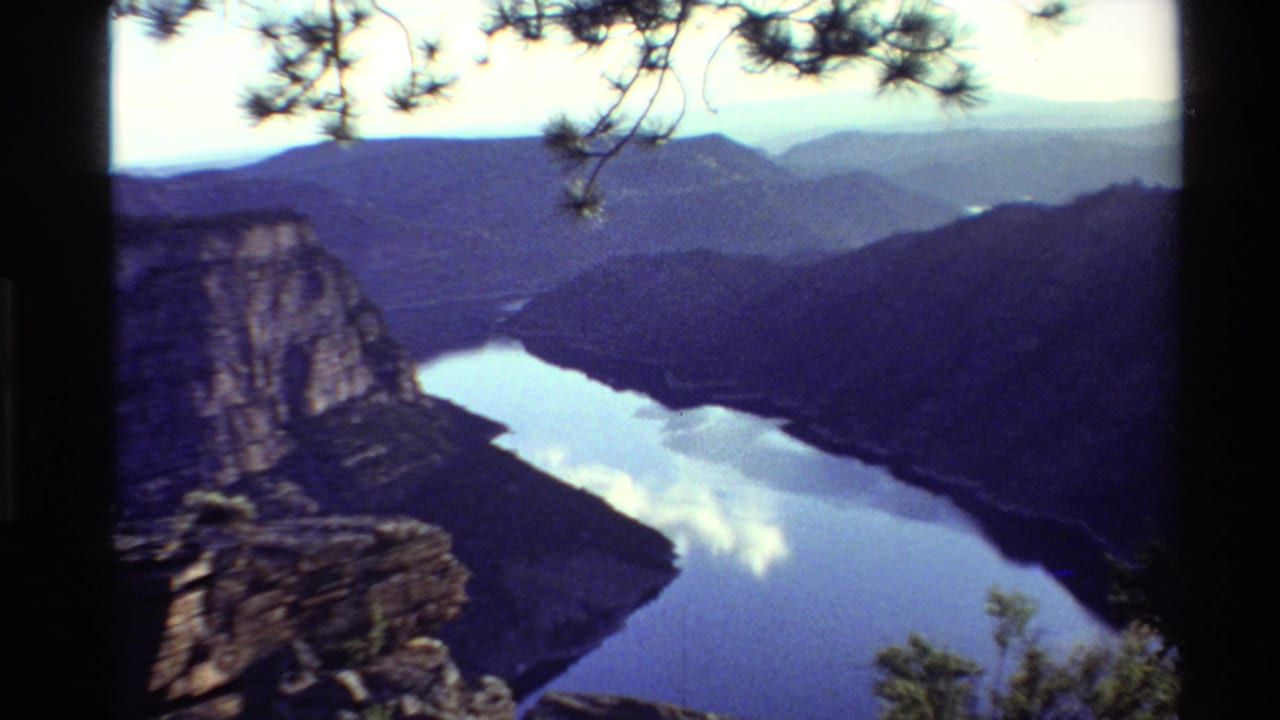 Canyon river view with a steep rock face, dark hills, pine branches, and reflected sky.