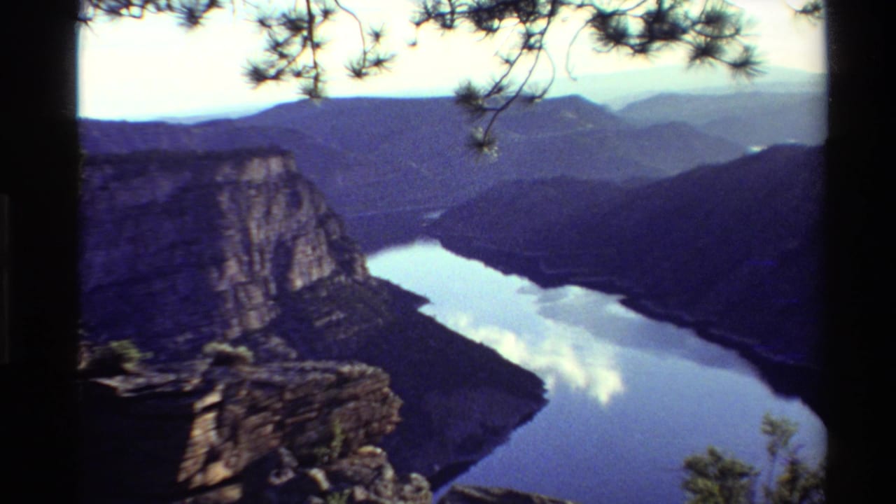 1980 Western USA 8mm frame showing a river bend below a rocky cliff, with wooded mountains and soft cloud reflections on the water.