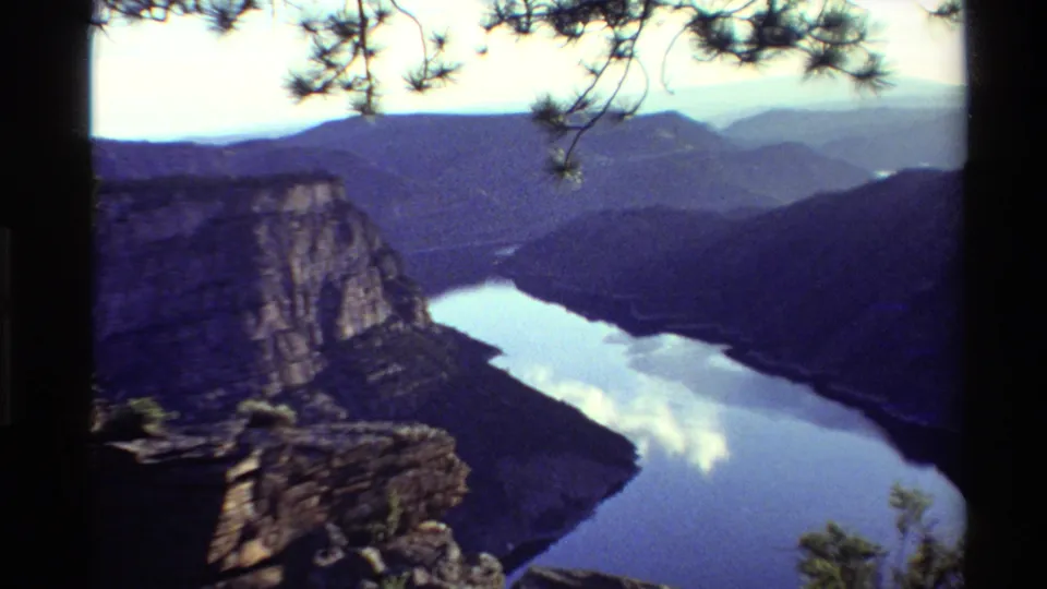 1980 Western USA 8mm frame showing a river bend below a rocky cliff, with wooded mountains and soft cloud reflections on the water.