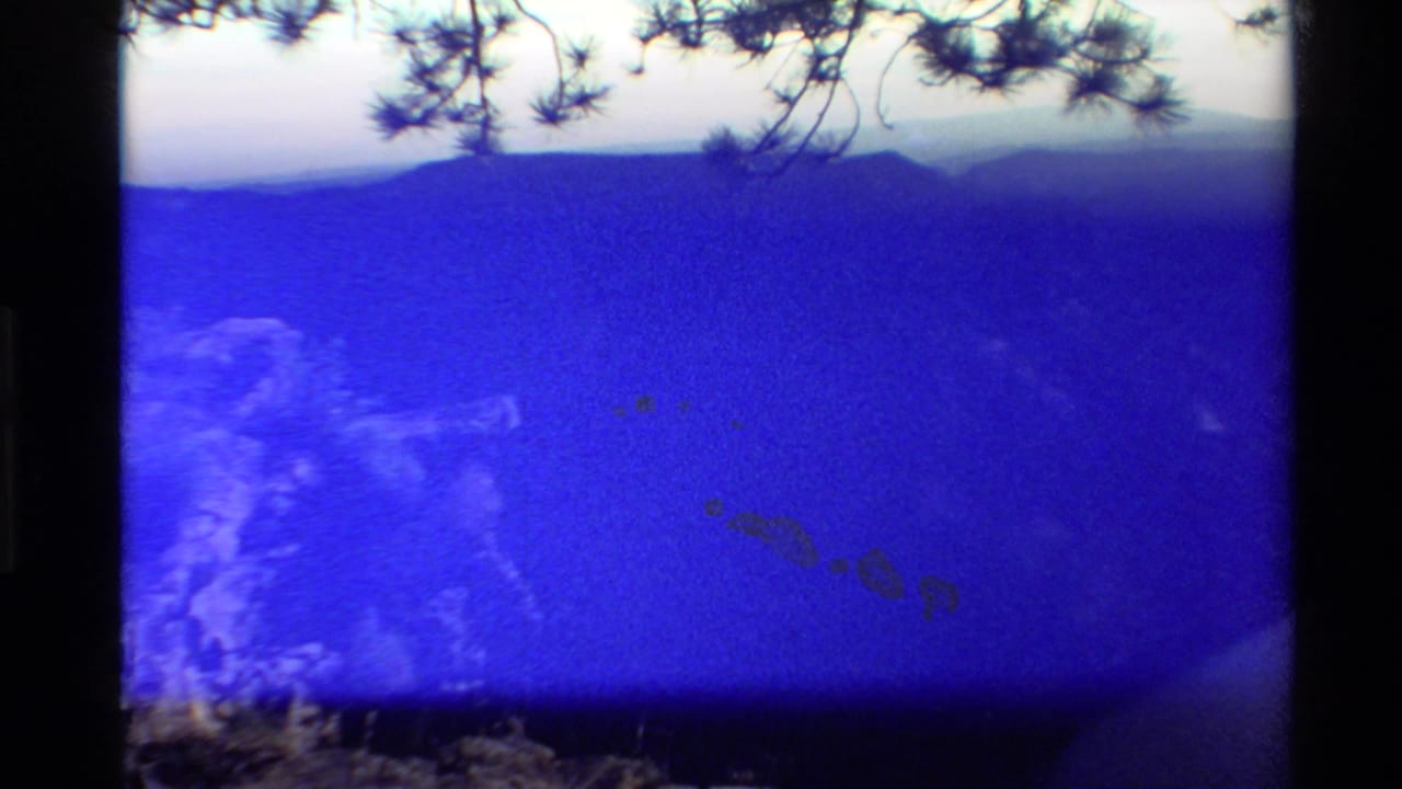 Strongly blue-shifted 1980 8mm frame with faint mountain ridges, reflective water, pine branches, and visible exposure variation.