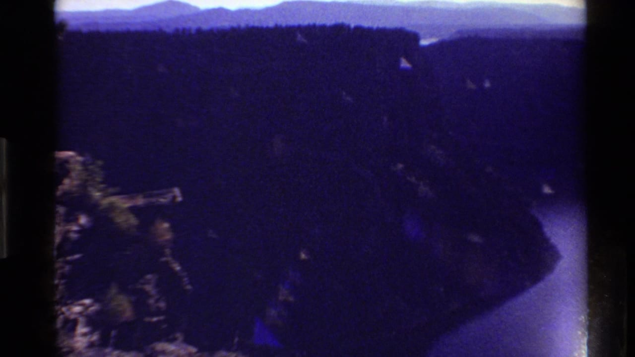 Reported 1980 Western USA home-movie frame showing a shadowed canyon, rocky edge, and curving river under blue-purple aged-film color.