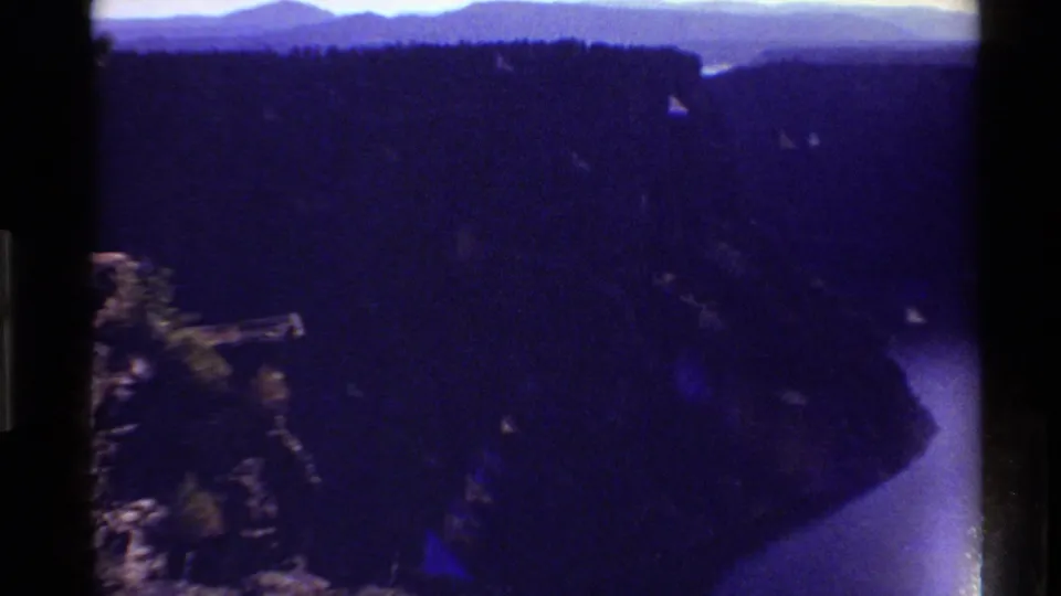 Reported 1980 Western USA home-movie frame showing a shadowed canyon, rocky edge, and curving river under blue-purple aged-film color.