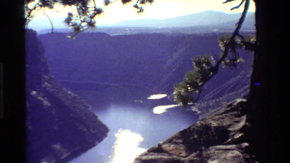 The final 8mm frame looks across a steep Western canyon toward river water, mountains, pine branches, and strong sun flare.