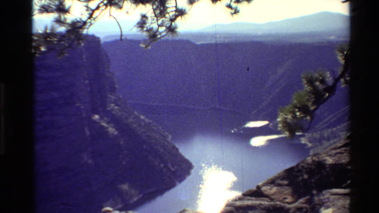 1980 Western USA 8mm still showing a canyon river from above, framed by pine needles, rock, and a hazy mountain horizon.