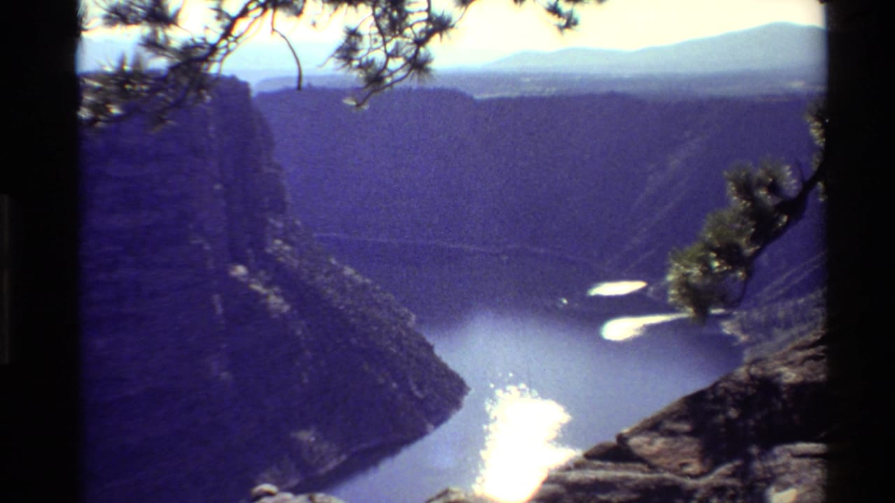 1980 Western USA landscape frame with a blue river in a deep canyon, foreground pine branches, and soft distant mountains under haze.
