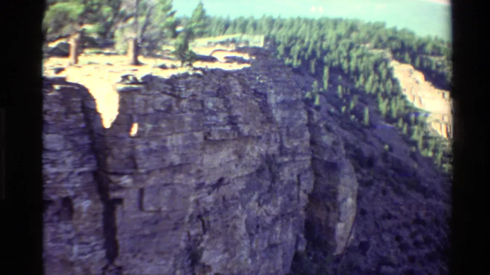 1980 8mm Western USA view across a massive cliff wall, showing rugged strata, rim trees, and forested terrain beyond.
