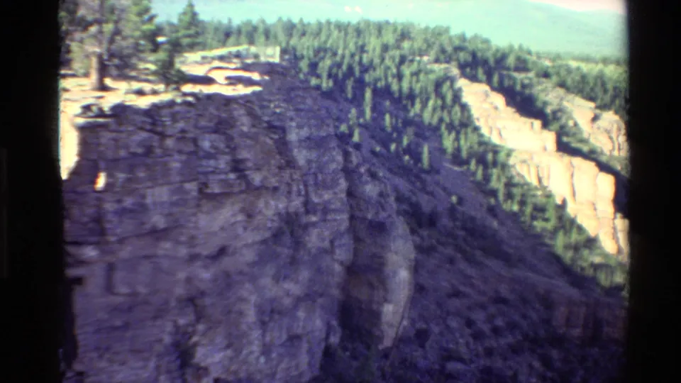 1980 8mm Western USA frame of a rugged cliff face beside a forested canyon rim, with soft focus and warm home-movie color.