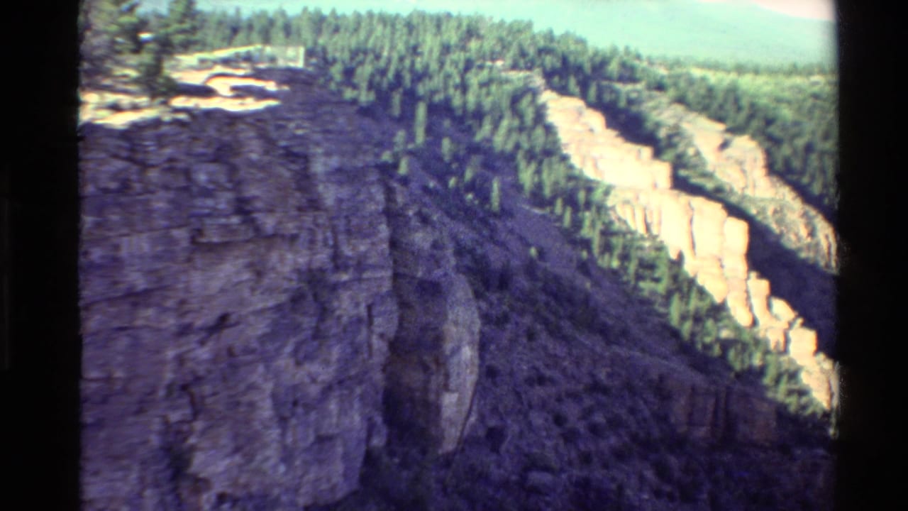 1980 Western U.S. 8mm frame showing a steep rocky cliff at left, evergreen forest above, and pale sunlit rock formations across a valley.