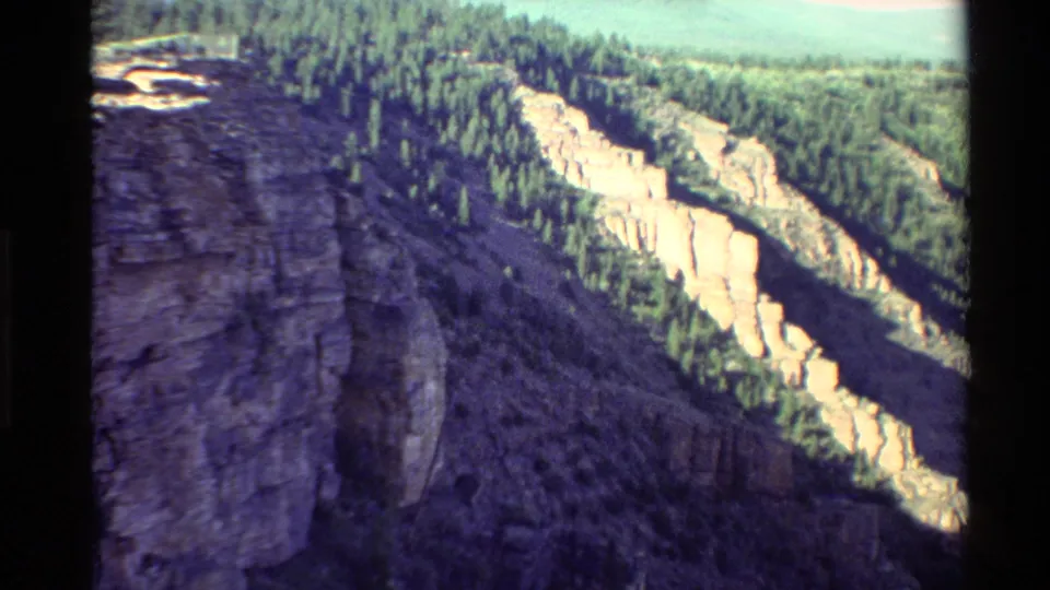 1980 Western U.S. 8mm frame with a broad rocky foreground wall, wooded upper slopes, and light-colored cliffs stretching across the middle distance.