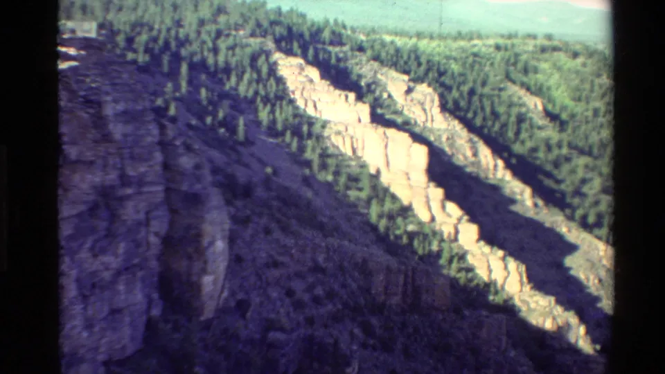 1980 Western U.S. 8mm frame showing pale cliff formations, dense evergreen cover, a dark foreground rock edge, and shadowed terrain below.