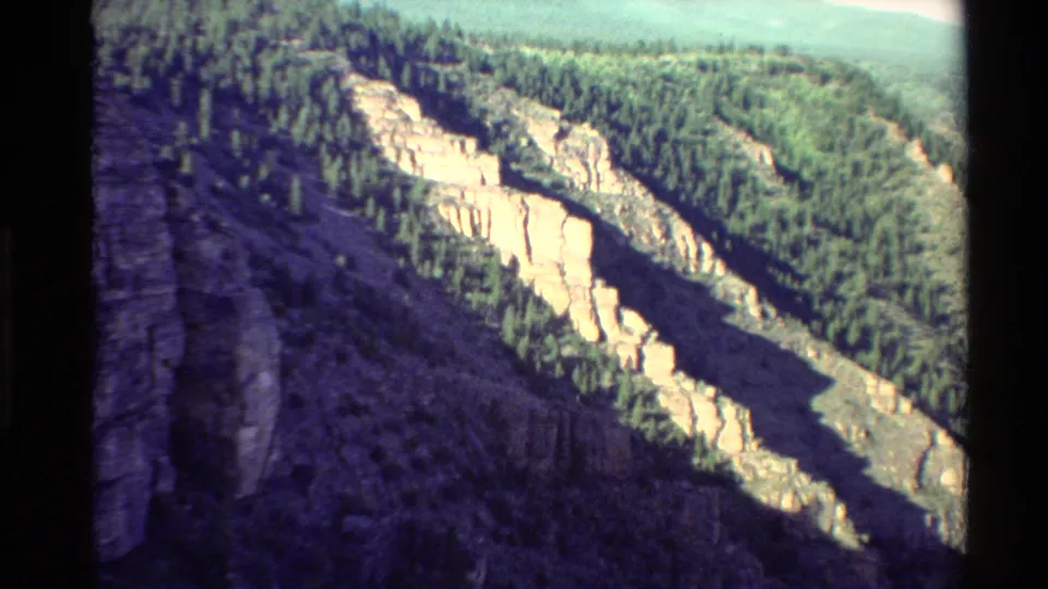 1980 Western U.S. 8mm frame with evergreen ridges, sunlit pale rock walls, dark valley shadows, and a partial rocky edge at left.