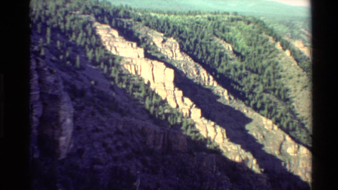 1980 8mm Western USA view of pale canyon cliffs, dark shadowed rock, and forested mountain slopes.
