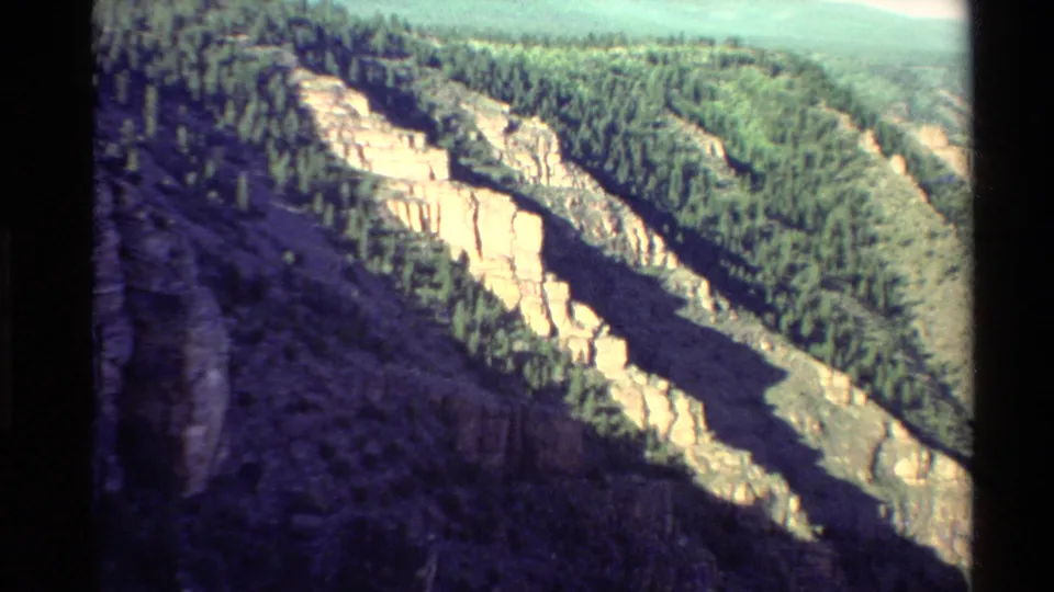 1980 8mm Western USA view of pale canyon cliffs, dark shadowed rock, and forested mountain slopes.