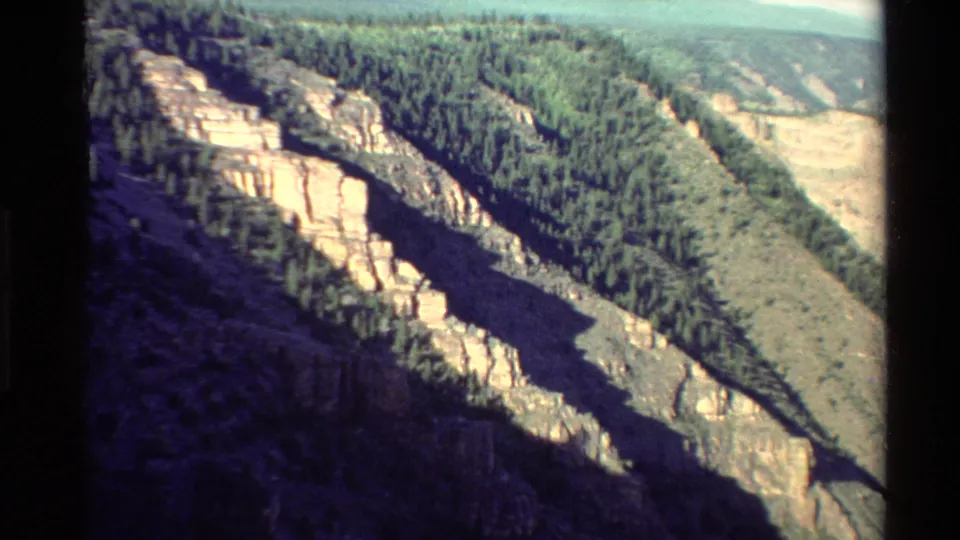 Vintage 8mm view of a rugged Western USA canyon, with sunlit cliffs, trees, and a shadowed drop below.