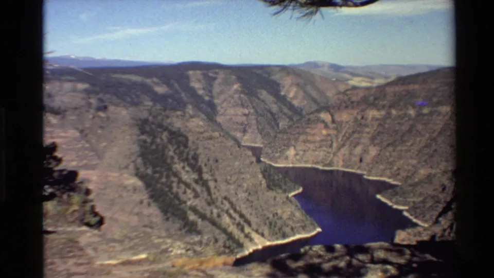 1980s Western USA home-movie frame showing a canyon river bend, dry rocky slopes, pines, and distant ridgelines.