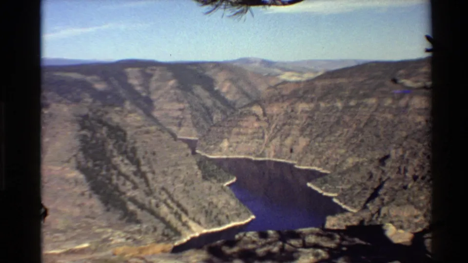 Western USA canyon vista on 8mm film, showing layered rocky slopes, a winding blue river or reservoir, and bright sky.