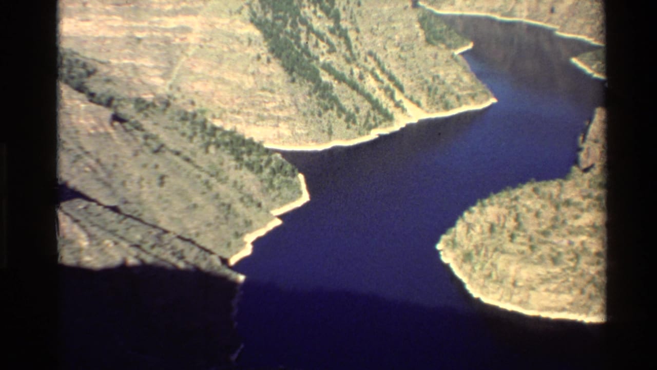 A vintage 8mm aerial view of a deep blue river curving between pale, rugged canyon slopes in the Western USA.