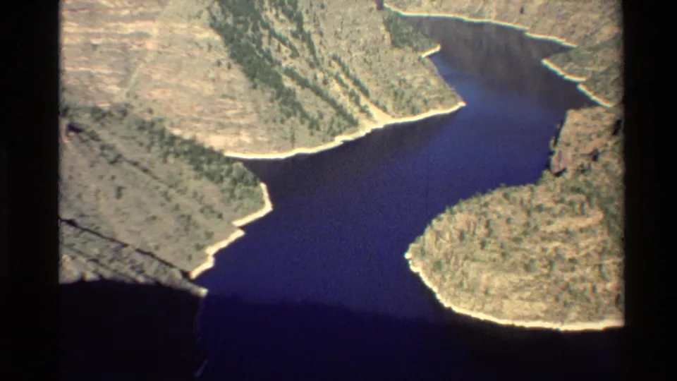 1980s 8mm aerial frame showing a blue river channel between steep canyon walls, dry slopes, and sparse tree-dotted terrain.