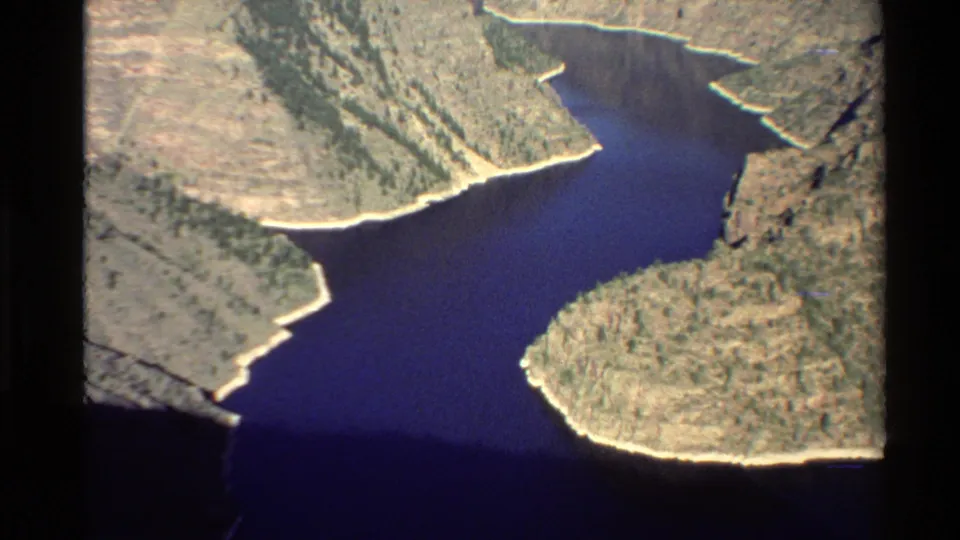 Vintage 8mm Western USA aerial view of blue water cutting through a rugged canyon with pale banks and sparse vegetation.