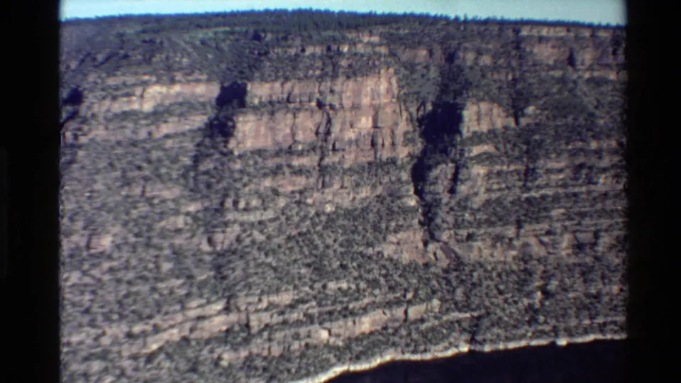 A striated canyon wall rises above dark river water in vintage 8mm footage, with layered rock and sparse vegetation visible on the cliff face.