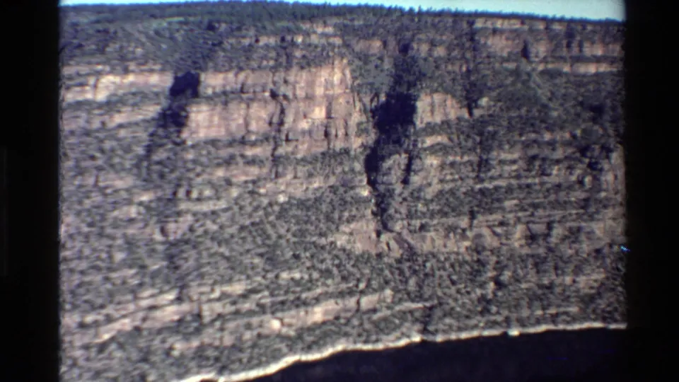Aerial 8mm frame of layered Western USA canyon cliffs with sparse vegetation and a dark river edge along the bottom of the scene.