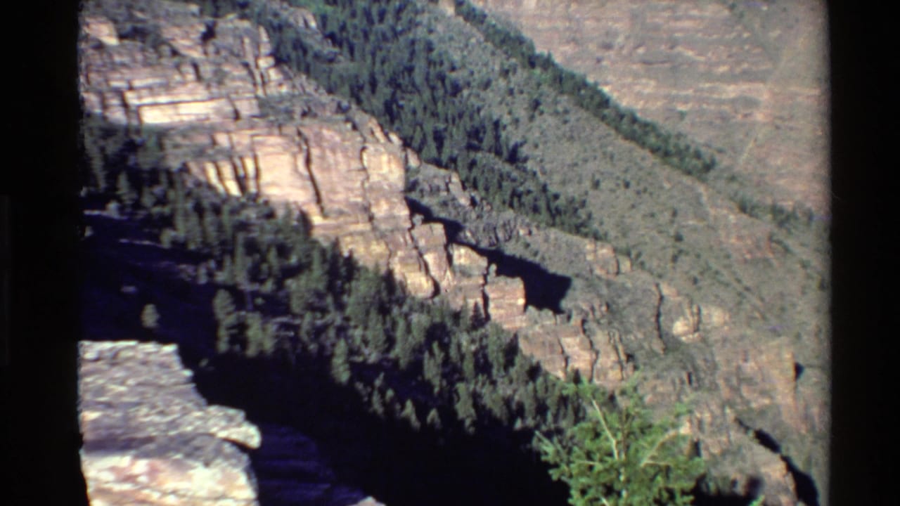 1980 Western U.S. 8mm frame showing layered canyon cliffs, evergreen trees, and a dark valley from an overlook perspective.