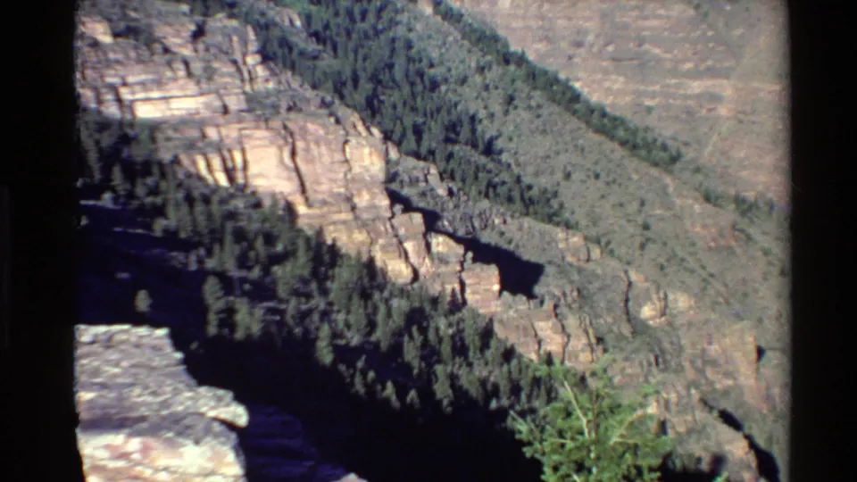 1980 Western U.S. 8mm frame showing layered canyon cliffs, evergreen trees, and a dark valley from an overlook perspective.