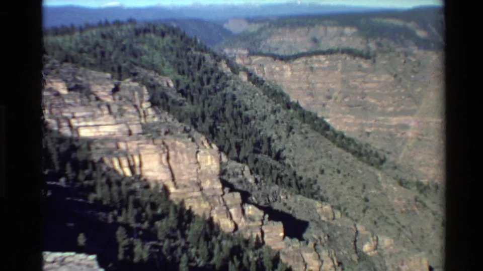 1980 Western U.S. 8mm frame showing a wider canyon overlook, with wooded slopes, rock formations, and distant mountains.