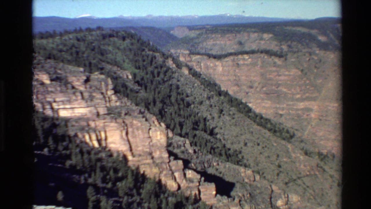 1980 8mm Western U.S. landscape view of a forested canyon rim, layered rock walls, distant ridges, and pale blue sky.