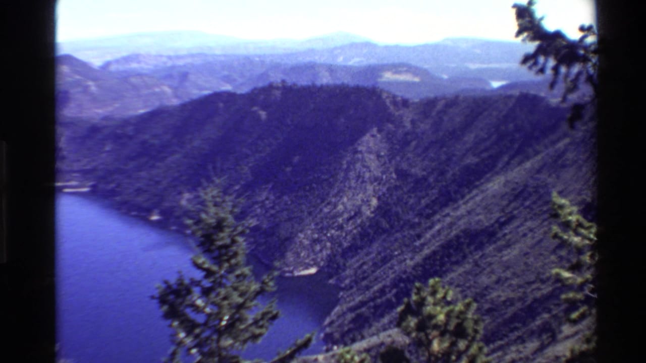 1980 8mm Western U.S. overlook view across blue water, steep forested slopes, distant ridges, and evergreen branches.