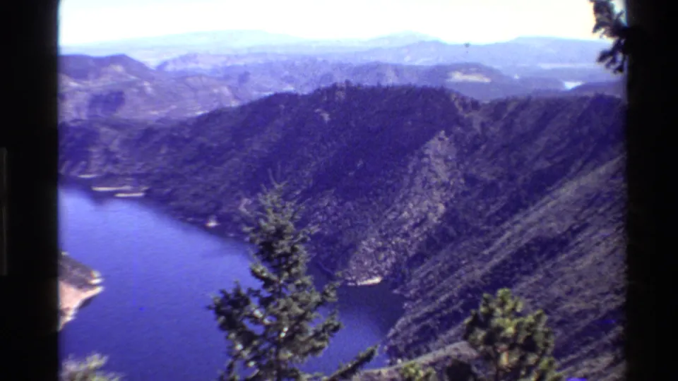 1980 8mm Western U.S. scenic view with a foreground evergreen, blue river or reservoir, steep mountainside, and distant ridgelines.