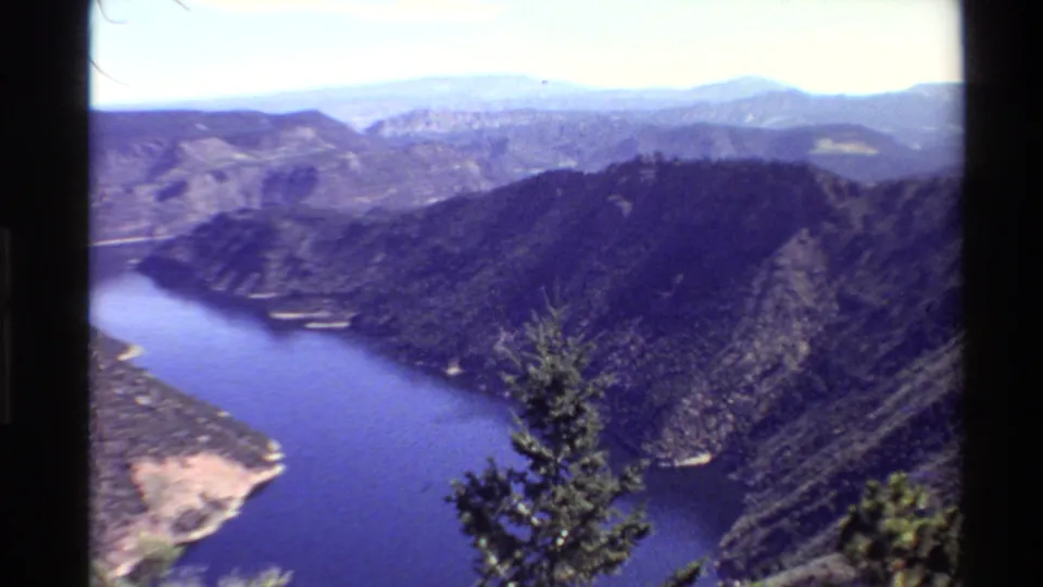 1980 8mm Western U.S. landscape frame showing blue water curving below a forested ridge, with distant hills and a small evergreen in front.