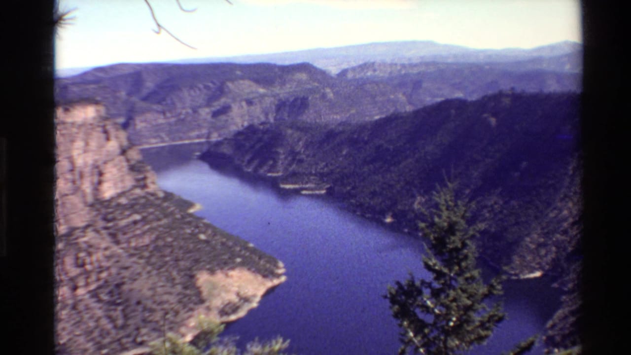 1980 Western U.S. home-movie frame showing a canyon lake, rocky cliff, wooded slopes, and evergreen branches from an overlook.