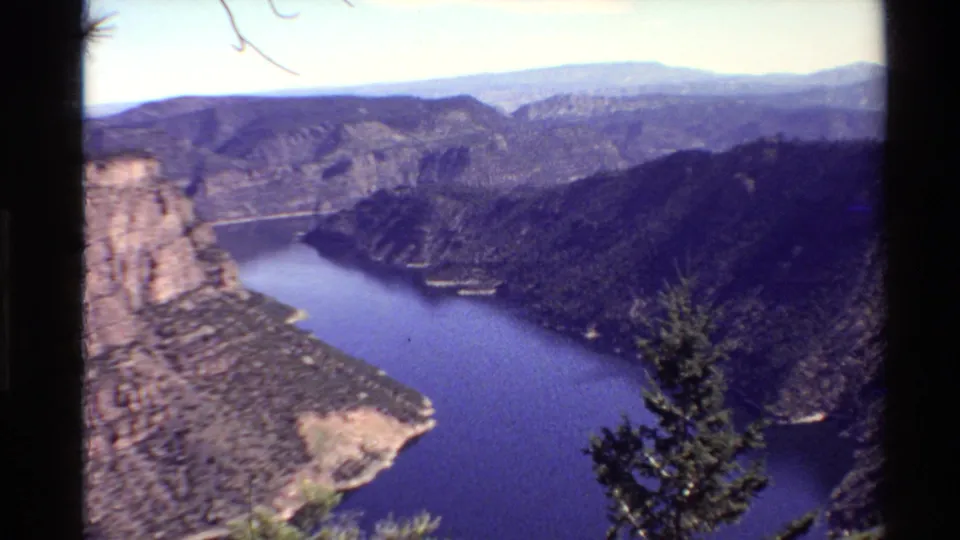 1980 Western U.S. home-movie frame showing a canyon lake, rocky cliff, wooded slopes, and evergreen branches from an overlook.