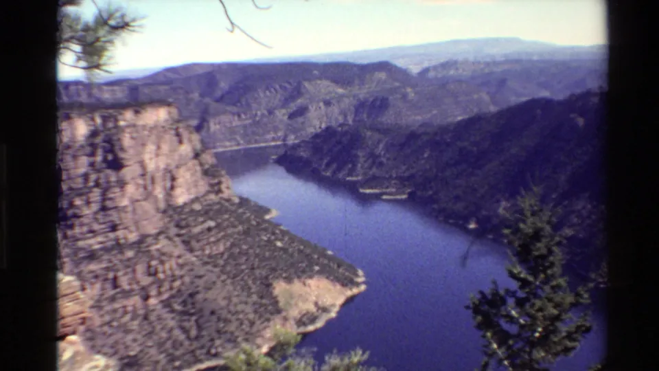 1980 Western U.S. 8mm frame with a prominent canyon wall, blue water below, forested terrain, and evergreen foreground.