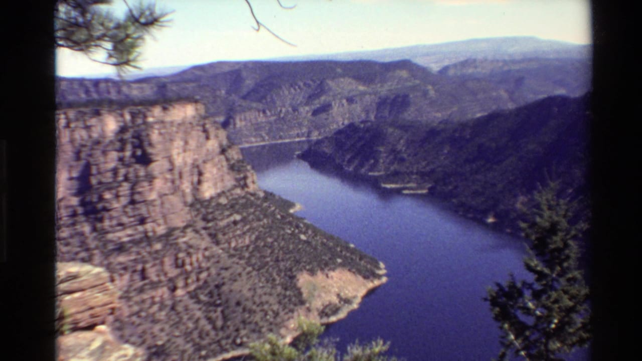1980 Western USA 8mm frame showing a canyon overlook with blue water, layered cliffs, distant ridges, and dark pine branches.