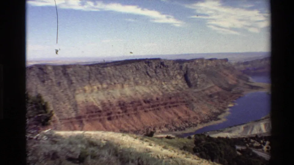 1980 Western USA 8mm frame showing a canyon rim, layered rock walls, blue water below, scrub vegetation, and pale sky.