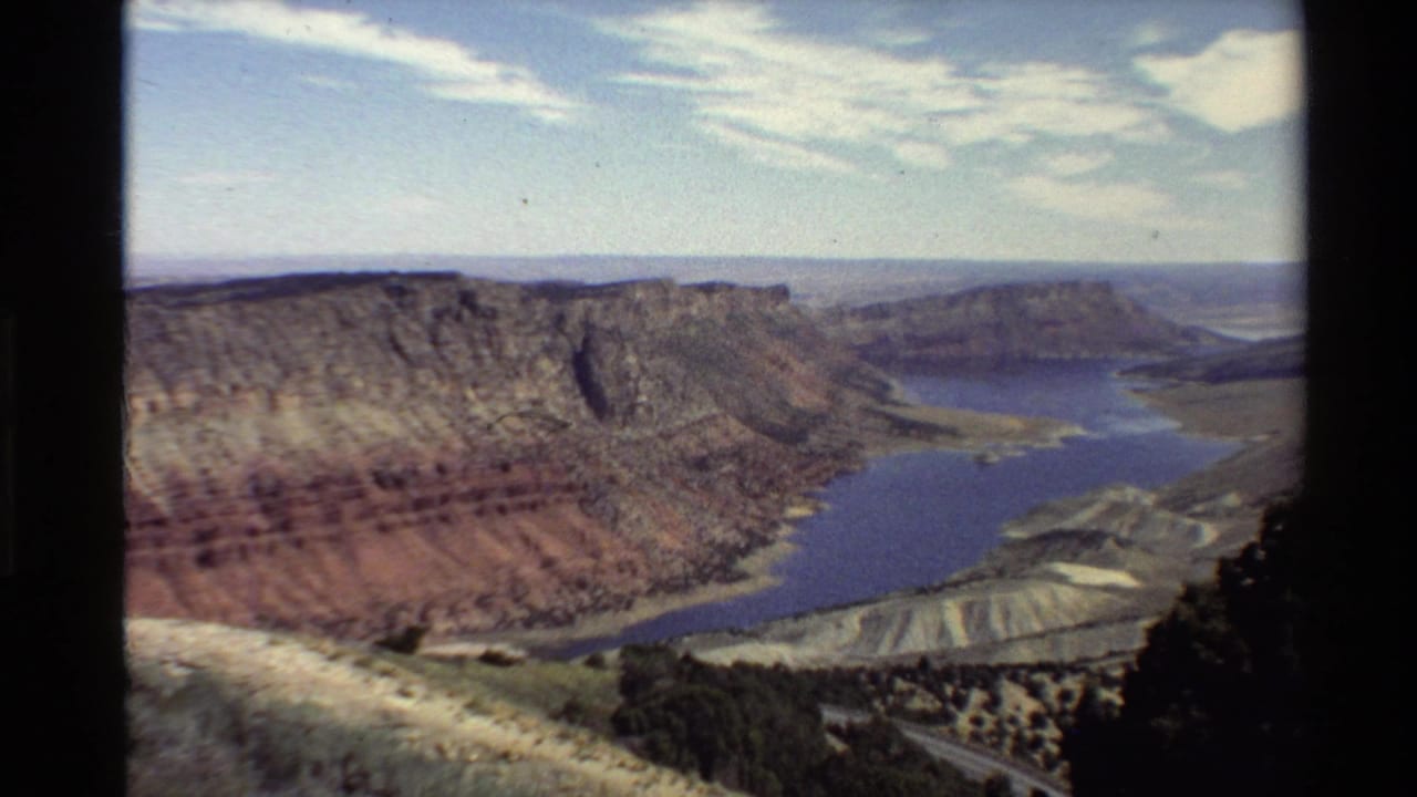 1980 archival home-movie frame of red-brown canyon formations, reservoir water, pale ridges, and a hazy horizon.