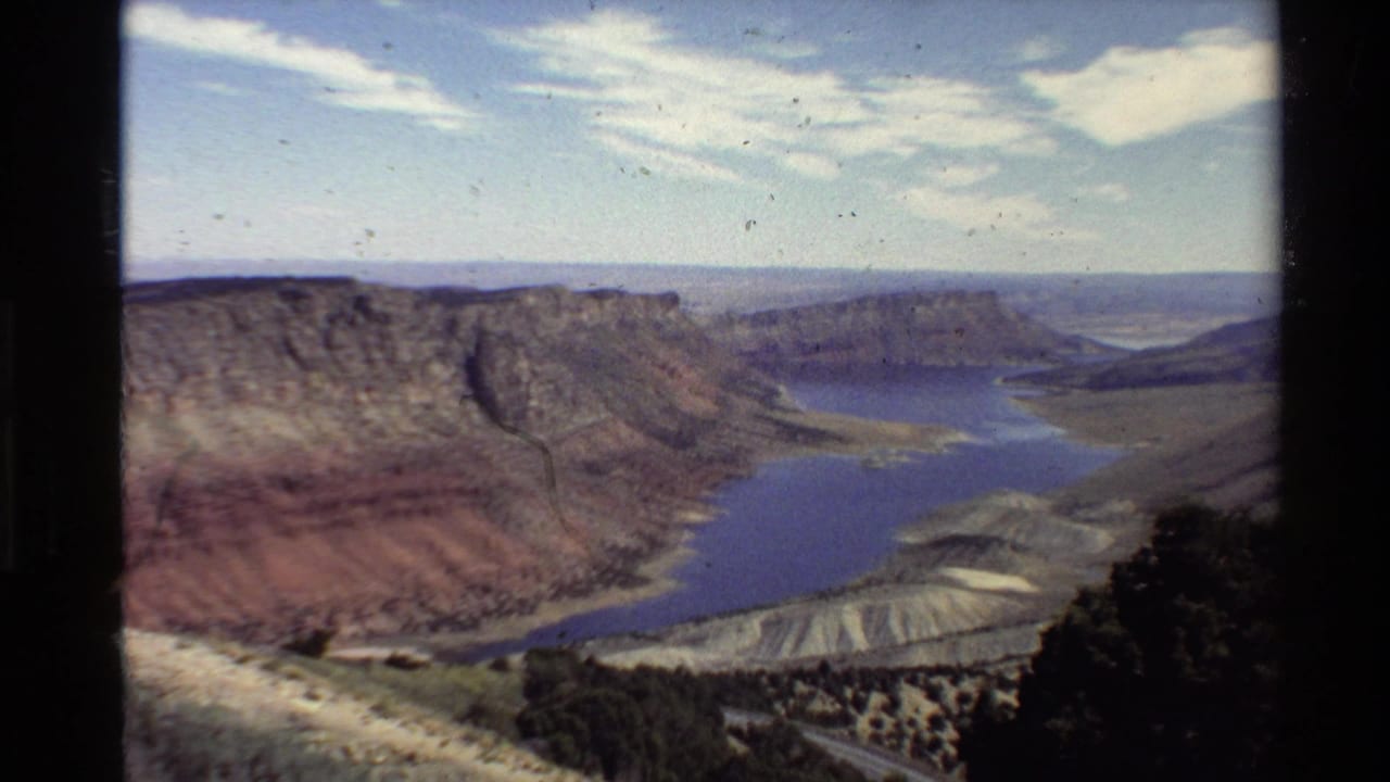 Western U.S. 8mm landscape frame showing canyon walls, a blue reservoir, cloud streaks, and visible archival film specks.