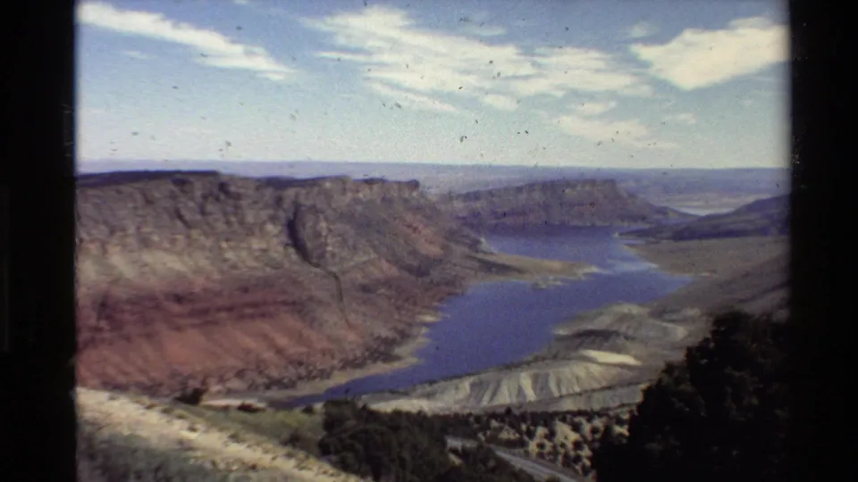 Western U.S. 8mm landscape frame showing canyon walls, a blue reservoir, cloud streaks, and visible archival film specks.