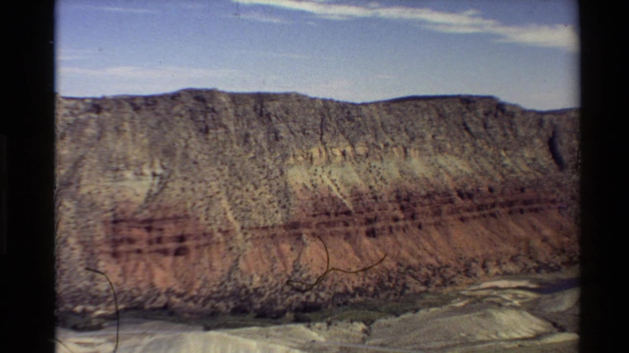 1980 8mm frame of a Western U.S. canyon wall seen from above, with red and tan strata, blue sky, and pale terrain below.