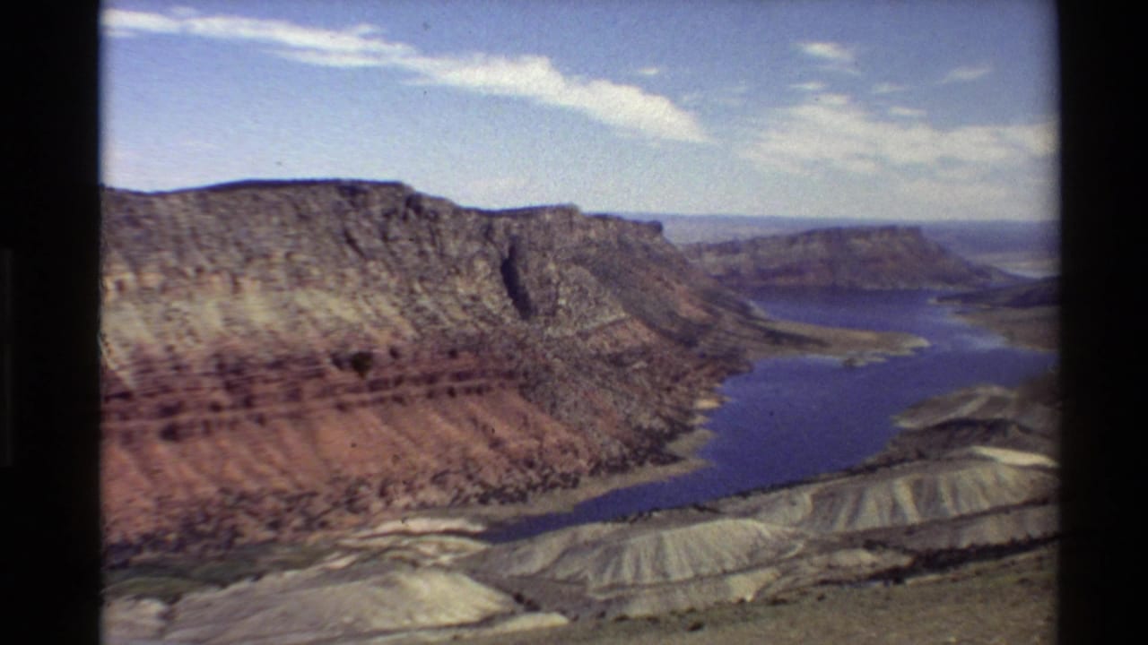 Wide 8mm aerial frame of a Western U.S. canyon, with a blue river, layered rock wall, pale slopes, and distant mesas under clouded sky.