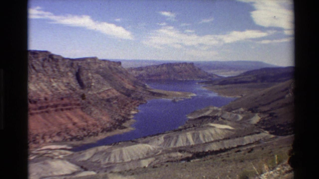 1980 Western U.S. 8mm view of a deep canyon, blue river, distant mesas, and scattered clouds from an elevated vantage point.