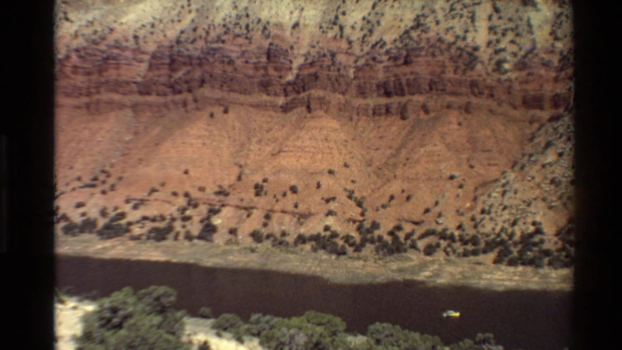 Elevated 8mm view of a layered red canyon wall above a dark river, with scrub vegetation and a small boat visible near the water.