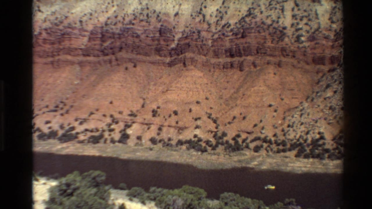 1980 Western U.S. canyon river scene with layered red rock, pale slopes, riverside vegetation, and a small boat on the water.