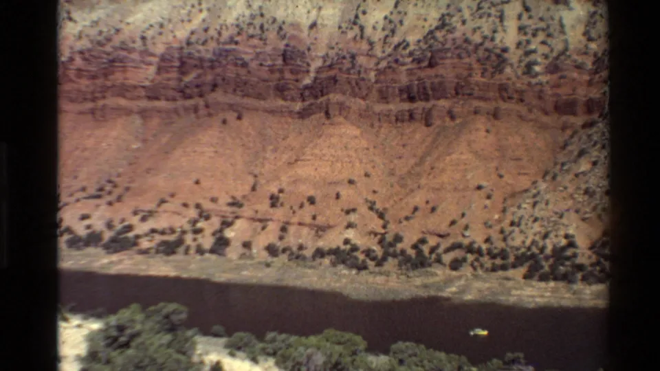 1980 Western U.S. canyon river scene with layered red rock, pale slopes, riverside vegetation, and a small boat on the water.