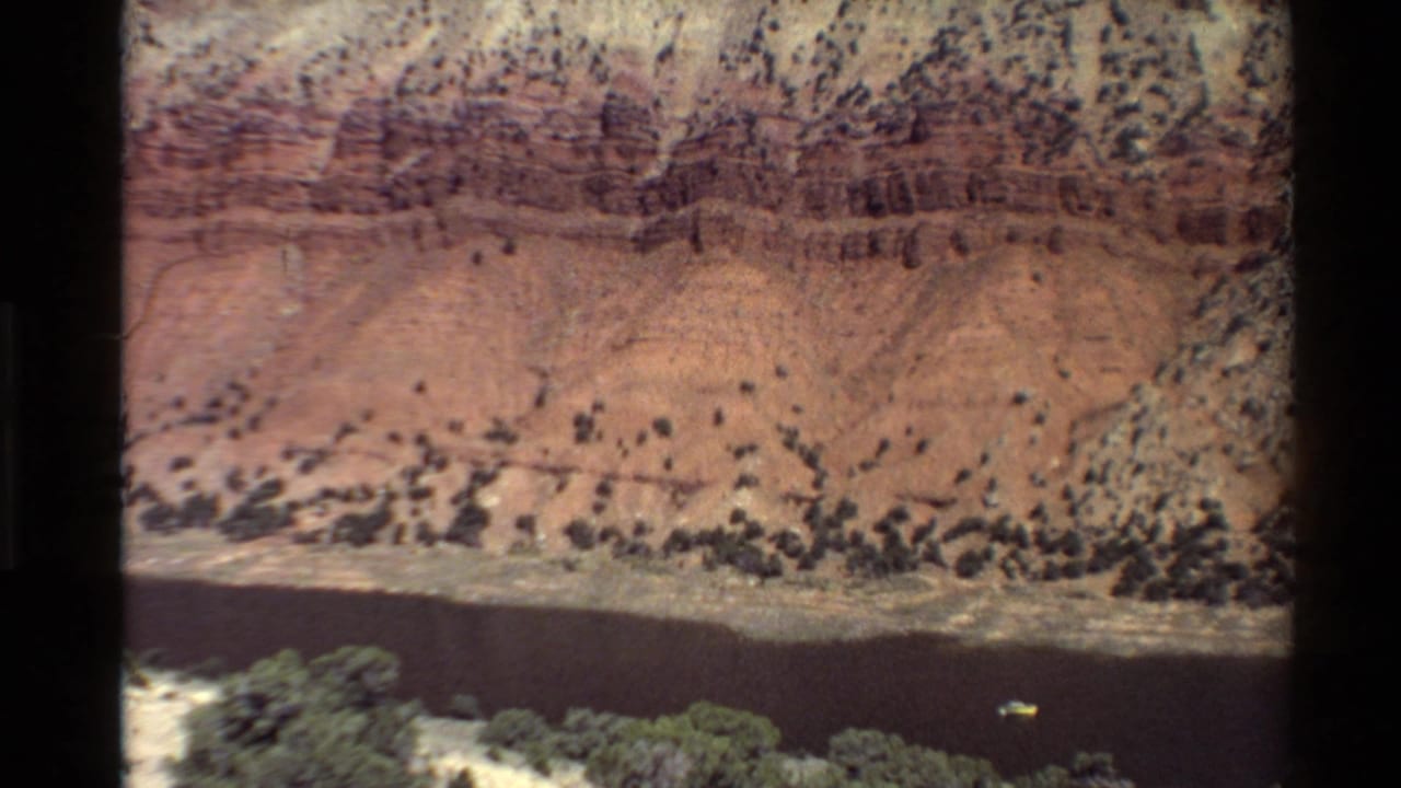 Soft, grainy 8mm frame of a Western U.S. canyon wall, dark river, and scrub vegetation from an elevated home-movie viewpoint.