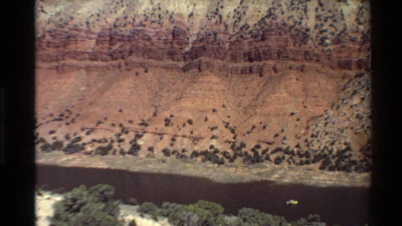 Red rock canyon wall above a dark river or reservoir with scrub vegetation in the foreground.